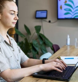 Woman sits in an office working on her laptop with a notebook next to her