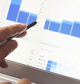 Photo of a hand with a pen pointing at graphs on a laptop screen