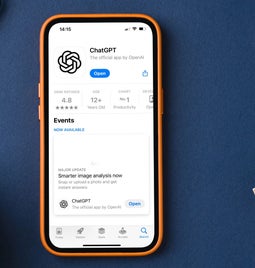 A smartphone displaying the ChatGPT app rating and features, surrounded by a coffee mug, glasses, and a notebook on a blue background.