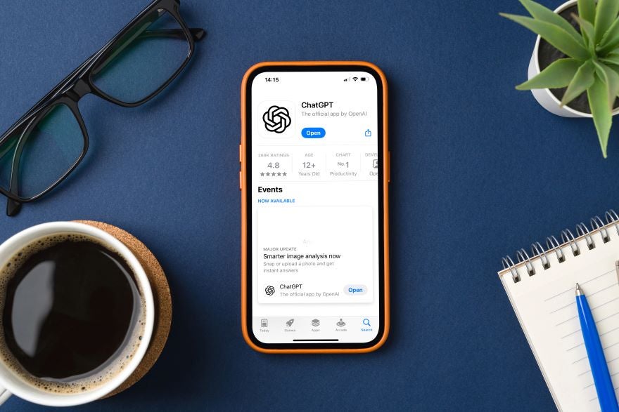 A smartphone displaying the ChatGPT app rating and features, surrounded by a coffee mug, glasses, and a notebook on a blue background.
