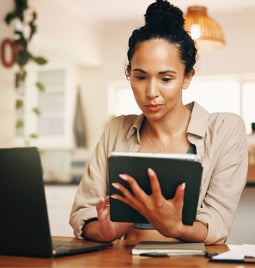 A person sits at a wooden table, holding a tablet, with a laptop and stationery in a cozy home office setting filled with plants.