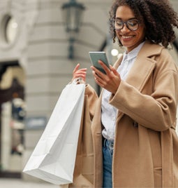 Woman in a coat standing outside with a couple of shopping bags while looking at her phone