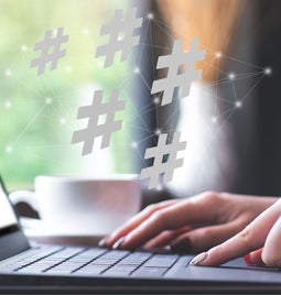 A person types on a laptop in a caf&eacute;, with floating hashtags above, symbolizing social media engagement. A cup is nearby.