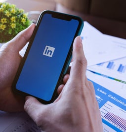 A person holds a smartphone displaying the LinkedIn logo, surrounded by papers with graphs and a small plant nearby.