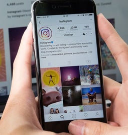 Hands hold a mobile phone showing Instagram's profile on Instagram, with a desktop behind also showing the same image