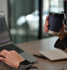 A person sat at a desk sipping coffee while editing code on a laptop