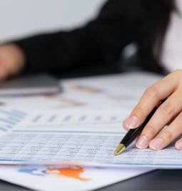 A closeup of paperwork on a desk and someone's hands pointing at a sheet with a pen in hand