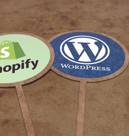 Two signs resting on the sand - one has the Shopify logo, while the other has the WordPress logo