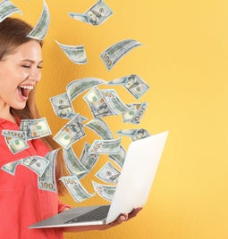 A person in a red shirt holds a laptop, surrounded by floating hundred-dollar bills against a bright yellow background.
