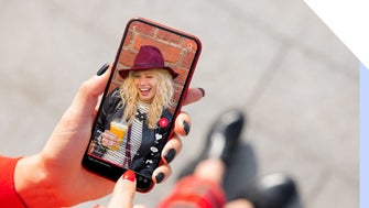 A hand holds a smartphone displaying a social media video, featuring a person with curly hair and a drink, against a brick background.