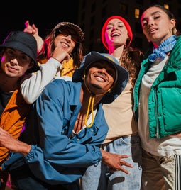 A group of five people in colorful streetwear poses playfully at night, with a vibrant pink light illuminating the background.