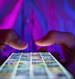 A person holding a smartphone illuminated by colorful lights, scrolling through a grid of images on the screen.