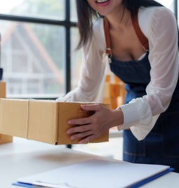 A woman in an apron lifts a cardboard box while preparing to record a video, surrounded by packing materials in a bright workspace.