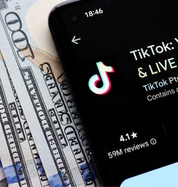 A smartphone displaying the TikTok app interface lies on a stack of hundred-dollar bills, highlighting digital media's connection to money.