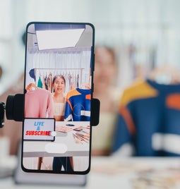 A smartphone on a tripod displays a clothing rack and makeup products in a bright studio, promoting a live streaming session.