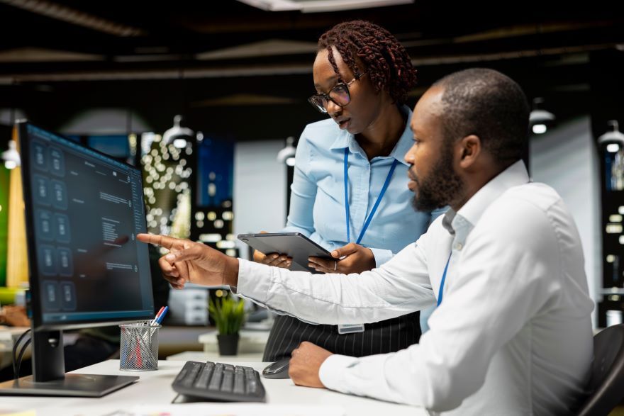 Two colleagues discuss a project at a modern office, focusing on a computer screen. One holds a tablet while pointing at the screen.