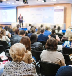 Conference hall people sat in rows of seats listening to a speaker on the stage