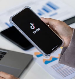 A phone with TikTok logo held above a table with a laptop on