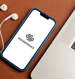 brown tables with white earphones, phone with squarespace's logo, and a laptop on it