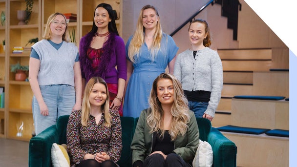 The Website Builder Expert team smiles at the camera, Heleana and Julia sat on a sofa and Emma, Holly, Lucy, and Annie stood behind them