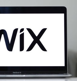 A close-up of a MacBook Air displaying the Wix logo on its screen, with a blurred background of bookshelves.